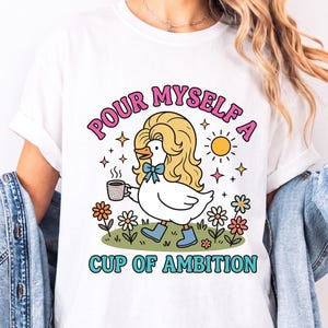 May include: A white t-shirt featuring a graphic of a white duck with long blonde hair, wearing blue boots and a blue bow tie, holding a coffee mug. The text reads "POUR MYSELF A CUP OF AMBITION" in pink and teal, surrounded by flowers and a sun.