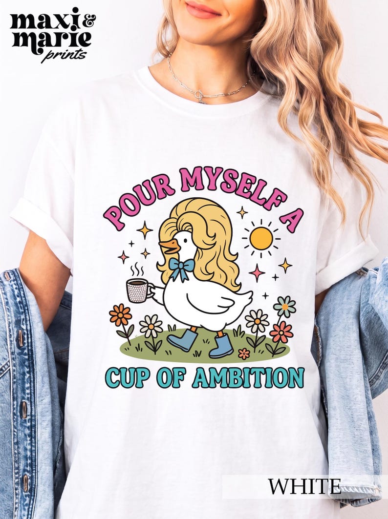 May include: A white t-shirt featuring a graphic of a white duck with long blonde hair, wearing blue boots and a blue bow tie, holding a coffee mug. The text reads "POUR MYSELF A CUP OF AMBITION" in pink and teal, surrounded by flowers and a sun.
