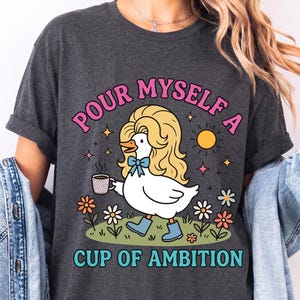 May include: A dark heather gray t-shirt featuring a graphic of a white duck with blonde hair, wearing blue boots and a blue bow, holding a coffee mug. The text reads "POUR MYSELF A CUP OF AMBITION" in pink and teal letters, surrounded by flowers and a sun.