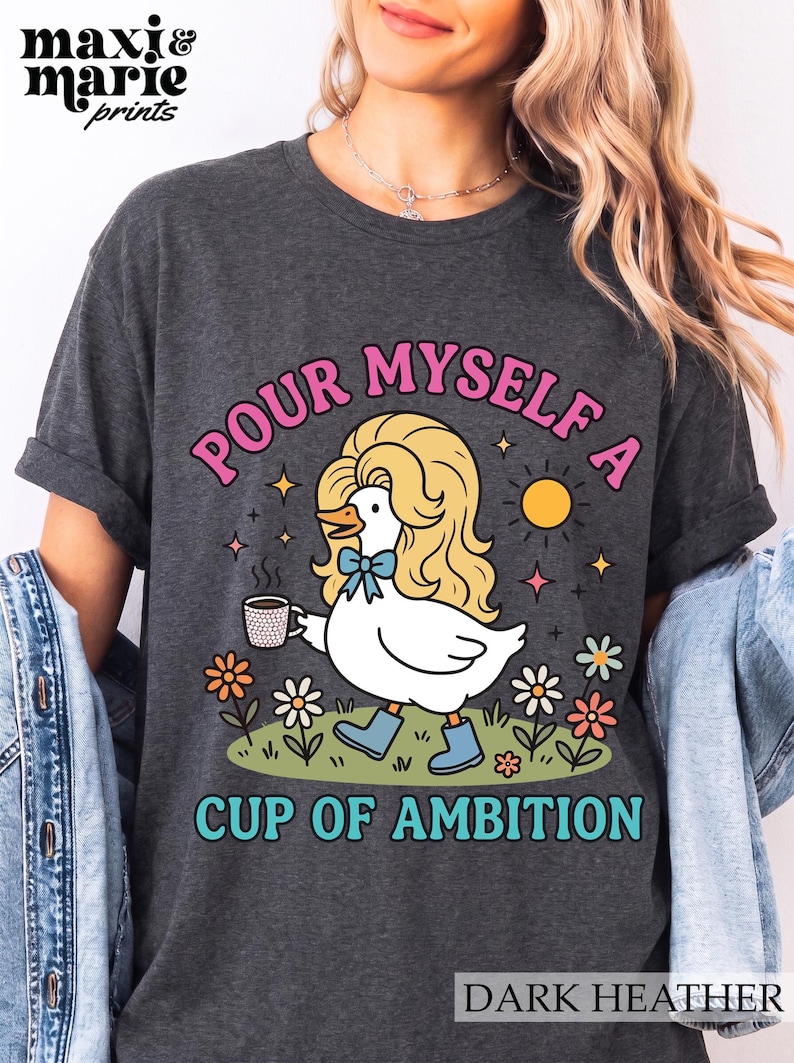 May include: A dark heather gray t-shirt featuring a graphic of a white duck with blonde hair, wearing blue boots and a blue bow, holding a coffee mug. The text reads "POUR MYSELF A CUP OF AMBITION" in pink and teal letters, surrounded by flowers and a sun.