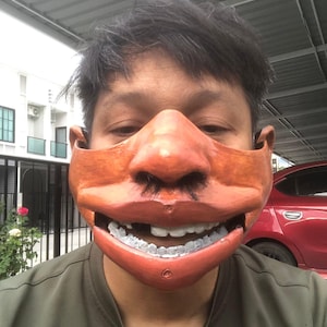 May include: A sculpted orange mask covering the lower face and nose, featuring a prominent nose with dark hair, a wide mouth with visible white teeth, and small circular indentations on the chin area, worn over a plain olive green t-shirt.