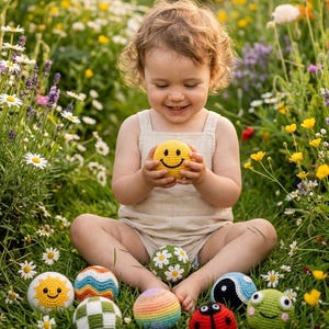 May include: A collection of small, round, crocheted yarn balls featuring various designs including a yellow smiley face, a sun, a ladybug, a green frog face, a yin-yang symbol, a rainbow gradient, a green and white checkerboard, and floral patterns.