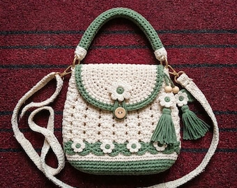 May include: A cream and sage green crochet handbag featuring a flap closure with a single wooden button, a top handle, and a detachable shoulder strap. The bag is decorated with small crocheted flower motifs and two green tassels.