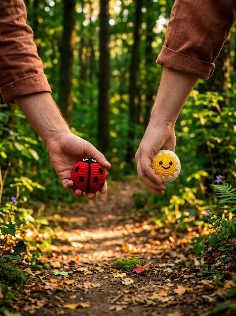 May include: Two round crochet toys, one a red ladybug with black spots and the other a yellow sun with a smiling face, held in palms against a blurred forest background with green foliage and fallen leaves on a dirt path.