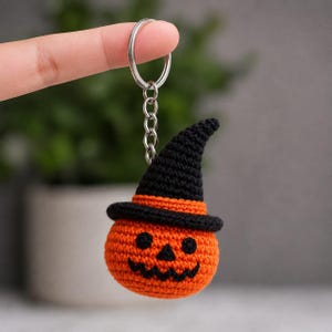 Op de afbeelding: Een gehaakte sleutelhanger in de vorm van een ronde oranje pompoen met een zwart gestikt jack-o'-lantern gezicht en een puntige zwarte heksenhoed. De pompoen is bevestigd aan een zilverkleurige metalen ring en een korte schakelketting.