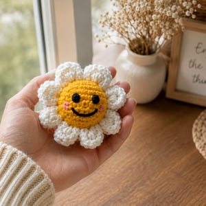 May include: A crocheted flower ornament featuring a bright yellow center with a black embroidered smiling face and pink cheeks, surrounded by white petals. The small, round, handcrafted textile item is held in a palm, showcasing its detailed yarn texture and circular shape.