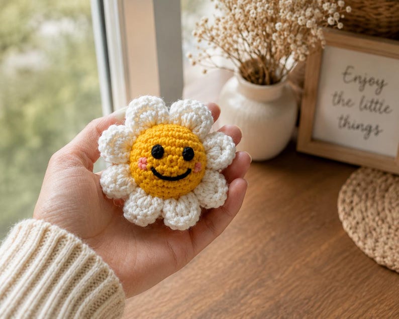 May include: A crocheted flower ornament featuring a bright yellow center with a black embroidered smiling face and pink cheeks, surrounded by white petals. The small, round, handcrafted textile item is held in a palm, showcasing its detailed yarn texture and circular shape.