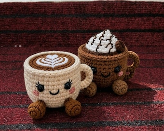 May include: Two crocheted coffee mugs with smiling faces, black bead eyes, and pink cheeks. The cream-colored mug features a white latte art heart design, while the brown mug is topped with white crocheted whipped cream and a small brown accent.