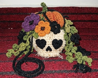 May include: A crocheted skull decoration in cream yarn with black heart-shaped eyes and a small triangular nose. It is adorned with a cluster of orange, purple, and black crocheted flowers, green vines, and a small orange pumpkin on top.