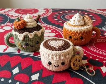May include: Three crocheted mugs with smiling faces. One is olive green with a pumpkin and cinnamon stick, one is orange with whipped cream and a heart, and one is cream-colored with a brown heart-shaped foam design and a small cookie charm.
