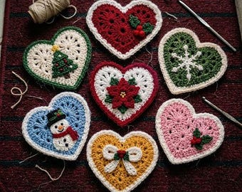May include: Seven crocheted heart-shaped ornaments featuring various designs including a Christmas tree, a snowman, a poinsettia, a snowflake, a bow, and holly berries, accompanied by a spool of yarn and two metal crochet hooks.