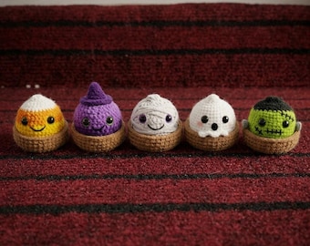 May include: Five small, round, crocheted figures sit in individual brown yarn bases. The figures include a candy corn, a purple witch, a white mummy, a white ghost, and a green monster with black stitches, all featuring black eyes.