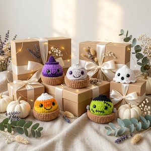 May include: A collection of six small crocheted Halloween figures including a purple witch, a white mummy, a white ghost, a candy corn, and a green monster, each sitting in a brown crocheted basket, arranged among gift boxes and white pumpkins.