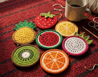 May include: A collection of seven round, crocheted fruit-themed coasters featuring designs of a strawberry, pineapple, watermelon, lemon, kiwi, orange, and dragon fruit, arranged alongside a ceramic mug and a ball of yarn.