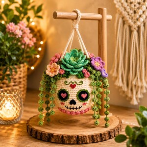 Op de afbeelding: Een gehaakte schedelplantenbak hangend aan een houten standaard, met een crèmekleurige basis, zwarte oogkassen met roze hartjes, een rood hartje op het voorhoofd en een gestikte mond. Het is gevuld met groene vetplanten en hangende ranken.