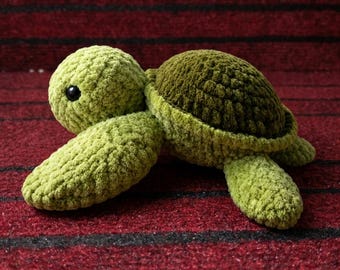 May include: A plush turtle toy crafted from soft, textured yarn. The head and flippers are a light lime green, while the rounded shell is a deep, dark olive green. The turtle features small, round, black plastic eyes.