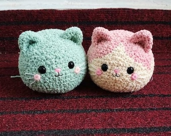May include: Two round crocheted cat plushies. The left cat is solid mint green with black eyes, a pink nose, and white whiskers. The right cat is pink and cream with black eyes, a pink nose, and white whiskers. Both have small ears.