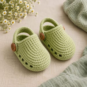 May include: A pair of light green crochet baby shoes featuring a rounded toe, a heel strap, and a small brown wooden button on each side. The shoes have a series of small rectangular cutouts along the sides and a textured, hand-stitched appearance.