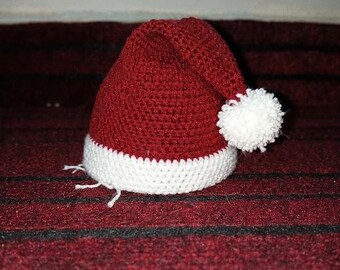 May include: A dark red crocheted hat with a white brim and a white pom-pom attached to the side. The hat features a dense, textured stitch pattern throughout the main body and a contrasting white band around the base with loose yarn ends.