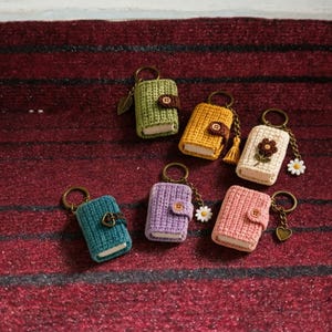 May include: Six miniature crochet book keychains in green, yellow, cream, teal, purple, and pink. Each features a button closure, a metal keyring, and unique charms including leaves, tassels, daisies, and hearts attached by small chains.