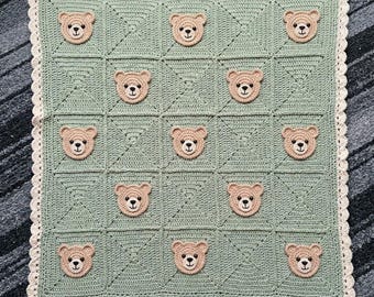May include: A square crochet blanket in sage green featuring a repeating grid pattern of eleven beige teddy bear faces. The blanket is finished with a scalloped cream-colored border around the entire edge, creating a textured design.