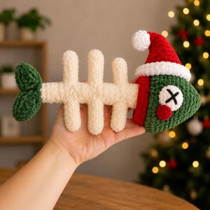 Pode incluir: Um brinquedo de pelúcia em crochê com formato de espinha de peixe, com coluna vertebral creme, cauda verde-escura e cabeça verde-escura com nariz vermelho e olhos em forma de X. O peixe usa um gorro de Papai Noel vermelho e branco.