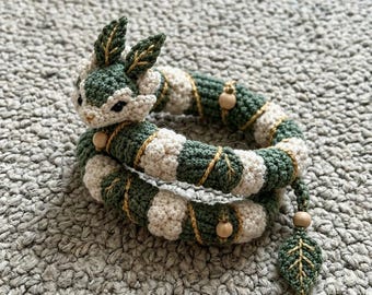 May include: A coiled crochet snake toy featuring alternating dark green and cream segments with gold thread accents. The head has pointed ears and dark eyes, while the tail ends in a green leaf-shaped charm. Small wooden beads are attached.