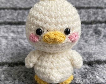 May include: A small, round, off-white crocheted duckling plush toy. It features a textured yarn surface, a small yellow beak, two black plastic bead eyes, and two pink embroidered circles on its cheeks. It has small yellow feet.