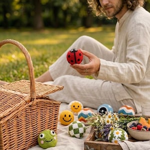 May include: A collection of small, round, crocheted toys featuring various designs including a red ladybug, a yellow smiley face, a green frog, a white and green checkered ball, a sun, a blue yin-yang, and a white ball with daisy flowers.