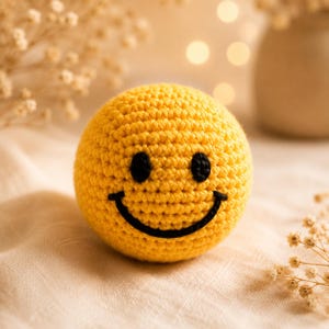 Handmade yellow smiley face crochet ball, soft sensory toy for babies, Montessori play, and cheerful nursery decor.
