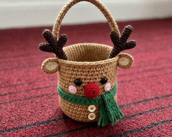 May include: A tan crochet basket featuring a reindeer design with dark brown antlers, small ears, black eyes, pink cheeks, and a red nose. The basket includes a green crocheted scarf and two white buttons on the front, with a single handle.