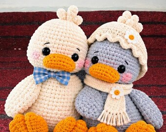 May include: Two crocheted duck plushies. The left duck is cream-colored with a blue and white checkered bowtie. The right duck is gray, wearing a cream-colored bonnet and scarf, each accented with a small white daisy flower applique.