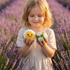 May include: Two round crochet balls. One ball is white with a yellow sun design featuring a smiling face. The second ball is green with a pattern of white daisies with yellow centers. Both items are spherical and textured with yarn stitches.