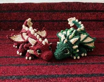 May include: Two crocheted dragon figurines, one in deep red and the other in forest green. Both creatures feature cream-colored wings, spines, and claws, with large, glossy black eyes and detailed textured yarn stitching throughout.