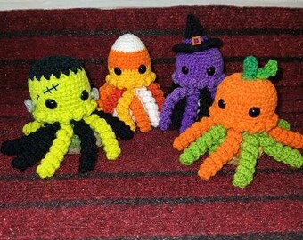 May include: Four crocheted octopus plushies featuring Halloween themes: a lime green monster with black hair, a white, orange, and yellow candy corn design, a purple octopus wearing a black witch hat, and an orange pumpkin with green leaves.
