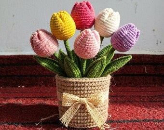 Puede incluir: Un ramo de tulipanes de ganchillo con seis flores en tonos rosa, amarillo, blanco y morado, con hojas verdes. Las flores están dispuestas en una maceta de ganchillo cilíndrica, texturizada y de color tostado, con un lazo.