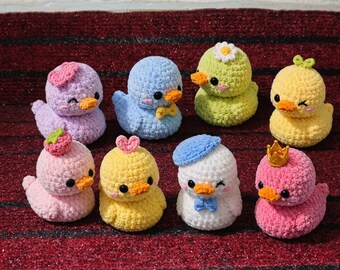 May include: A collection of eight small, crocheted duck toys in various colors including purple, blue, lime green, yellow, pink, and white. Each duck features black bead eyes, an orange beak, and unique accessories like hats, bows, or crowns.