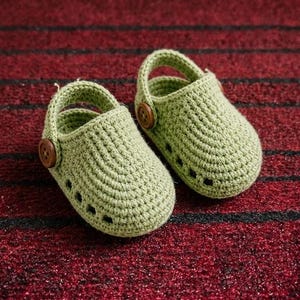May include: A pair of light green crochet baby shoes featuring a rounded toe, a heel strap, and a brown wooden button on each side. The shoes have a series of small rectangular cutouts along the sides, mimicking the design of a classic clog.