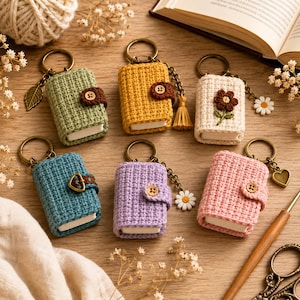 Könnte beinhalten: Sechs gehäkelte Miniatur-Buch-Schlüsselanhänger in Grün, Gelb, Creme, Blau, Lila und Pink. Jeder Schlüsselanhänger verfügt über einen Metallring, einen kleinen Knopf- oder Schnallenverschluss sowie dekorative Anhänger wie Blatt, Quaste, Gänseblümchen oder Herz.