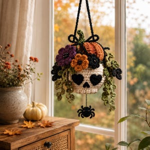 Op de afbeelding: Een gehaakte hangdecoratie met een crèmekleurige schedel met zwarte hartvormige ogen, bekroond met oranje en paarse bloemen, groene bladeren en een kleine pompoen. Een zwarte gehaakte spin hangt aan de onderkant van de schedel.