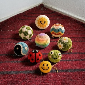 May include: A collection of nine spherical crochet ornaments featuring various designs including a yellow sun, a green and white checkerboard, a rainbow gradient, a yin-yang symbol, a ladybug, a green frog, a smiley face, and floral patterns.