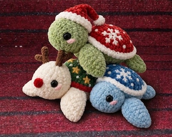 May include: Three crocheted plush turtles stacked together. The top turtle is light green with a red Santa hat and a red shell featuring white snowflakes. The bottom left turtle is white with brown antlers and a red nose. The bottom right turtle is light blue with a blue shell featuring white snowflakes.