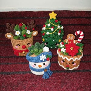 Könnte beinhalten: Vier gehäkelte Pflanzentopfhüllen mit weihnachtlichen Motiven: ein braunes Rentier mit Geweih, ein grüner Weihnachtsbaum mit gelbem Stern, ein blau-weißer Schneemann mit Schal und ein brauner Lebkuchenmann mit einer Zuckerstange.