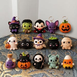 May include: A collection of fifteen small, crocheted Halloween-themed keychains featuring various characters including a black cat, Frankenstein's monster, a vampire, a bat, a pumpkin, a ghost, a spider, an owl, a cauldron, a mummy, a potion bottle, a cat in a pumpkin, a grim reaper, a zombie, and candy corn.