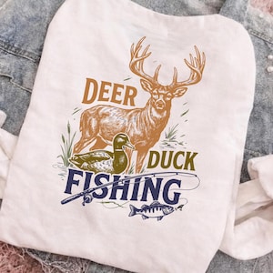 May include: A white t-shirt featuring a graphic print of a brown deer, a green duck, and a blue fishing rod with a fish. The text DEER, DUCK, and FISHING is printed in bold, coloured lettering around the illustrated wildlife and fishing gear.