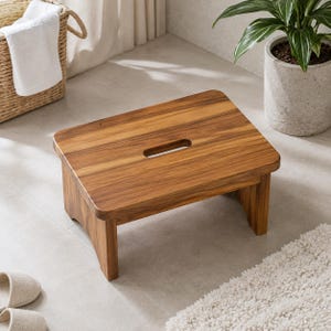 Puede incluir: Un taburete rectangular de madera con un acabado de veta natural en color marrón. El taburete presenta una superficie superior lisa y plana con un asa central recortada en forma de óvalo alargado para facilitar su transporte y cuatro patas integradas.