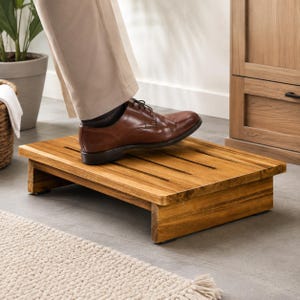 May include: A rectangular wooden footrest featuring a slatted top surface with a natural brown wood grain finish. The low-profile platform is supported by two solid side panels, providing a stable base for resting feet while seated in a chair.