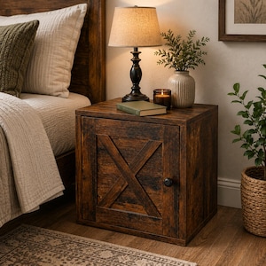 May include: A square wooden nightstand with a dark brown finish and a single cabinet door featuring a prominent X-shaped brace design. The cabinet has a small black round knob and sits beside a bed, supporting a lamp, a book, and a candle.