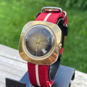 May include: A Timex wristwatch with a gold-tone square case and a round, dark brown gradient dial. The watch features gold-tone hands, a date window at the four o'clock position, and a red nylon strap with a central white vertical stripe.