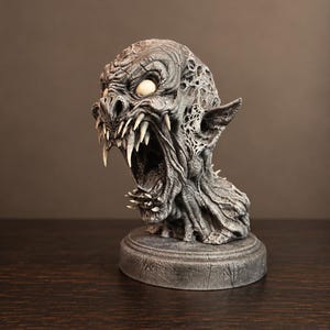 Pode incluir: Um busto de monstro cinzento e texturizado com a boca aberta, dentes afiados, orelhas proeminentes e olhos brancos sólidos. A criatura tem uma pele enrugada e desgastada, montada sobre uma base circular cinzenta a condizer.